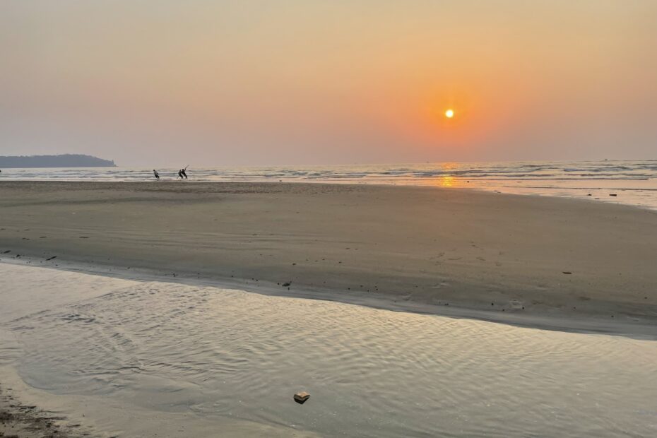 The sun setting through a hazy sky over a sandy beach in Goa, India.